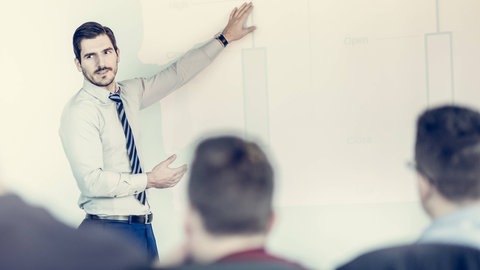 Ein Mann erklärt während einer Präsentation: Der Begriff "Powerpoint-Genau" stammt von dem Freiburger Linguisten Peter Auer. Es ist ein junges sprachliches Phänomen, das häufig in Vorträgen oder Prüfungssituationen auftaucht. 