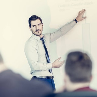 Ein Mann erklärt während einer Präsentation: Der Begriff "Powerpoint-Genau" stammt von dem Freiburger Linguisten Peter Auer. Es ist ein junges sprachliches Phänomen, das häufig in Vorträgen oder Prüfungssituationen auftaucht. 
