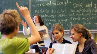 Vor einer mit lateinischen Sprichwörtern beschriebenen Tafel erteilt eine Lehrerin Lateinunterricht. Schülerinnen und Schüler arbeiten in einer Gruppe: Das Schulfach Latein hat nicht den besten Ruf. Doch die alte Sprache bastelt an einer neuen Zukunft. Weniger elitär, mehr Geschichte und Alltag. Welches Potenzial steckt in Latein?