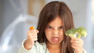 Kleines Mädchen schneidet wilde Grimasse und hält Brokkoli und Möhre von sich: Wie etwas schmeckt, legen nicht nur Zunge und Nase fest. Auch unsere Erfahrungen bestimmen darüber, vor allem aber die Kultur, in der wir leben. 