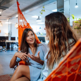 Zwei junge Frauen unterhalten sich in einer Hängematte in einem modernen Büro: Viele der um die Jahrtausendwende Geborenen würden gerne gleich mit einem Teilzeitjob starten. Studien zeigen auch, dass junge Menschen der sogenannten Gen Z Arbeit und Freizeit voneinander trennen und lieber im Büro als im Homeoffice arbeiten möchten. 