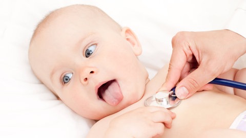 Kleinkind wird mit Stethoskop untersucht und streckt die Zunge heraus: Seit längerem ist der Notstand in der Pädiatrie bekannt, die Gründe dafür: zu wenige Fachkräfte, zu viel Bürokratie, zu geringe Honorierung, immer mehr Vorsorgeuntersuchungen, Engpässe bei Medikamenten, aber auch ein Trend bei Eltern zu mehr Arztbesuchen. 