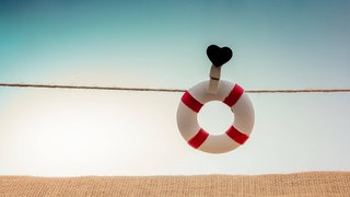 An einer Schnur befestigter Rettungsring mit schwarzem Herzsymbol: Mehr als 9.000 Menschen nehmen sich jedes Jahr in Deutschland das Leben. Forscherinnen und Forscher haben wirksame Therapien, Apps und Beratungskonzepte für Betroffene und Angehörige mit Suizidgedanken entwickelt. Allerdings fehlt Geld, um die Hilfsangebote überall verfügbar zu machen. 