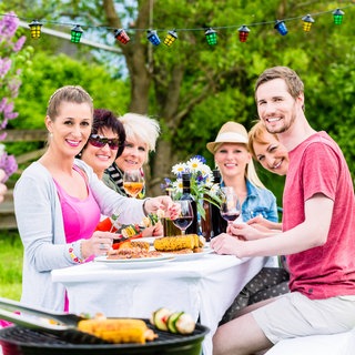 Grillfest im Garten: Sechs Menschen sitzen am Tisch, eine Person grillt: Einerseits werden Nachbarn immer zwischen Anonymität, Konflikt, Freundschaft und Gemeinschaft pendeln. Andererseits leben einige – auch mithilfe moderner Stadtplanung – inzwischen vor, was sie in guter Nachbarschaft gewinnen können.