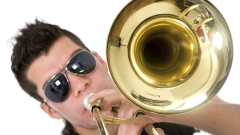 Junger Mann mit Sonnenbrille bläst in eine Posaune: Die Wucht von Trompete, Posaune und Tuba, moderne Rhythmen und Elemente von Techno bis Pop – neue Blasmusik-Combos sind mitreißend.