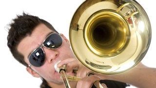 Junger Mann mit Sonnenbrille bläst in eine Posaune: Die Wucht von Trompete, Posaune und Tuba, moderne Rhythmen und Elemente von Techno bis Pop – neue Blasmusik-Combos sind mitreißend.