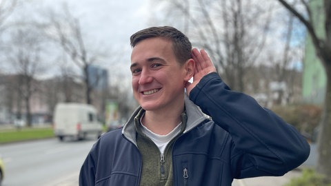 Benedict Kolajka hat selbst eine Hörschädigung und hält lachend eine Hand hinters Ohr: Schlecht hören heißt oft: nicht dazugehören. Mit Schwerhörigkeit steigt das Risiko für Isolation, Depression und Demenz. Trotzdem überspielen viele Betroffene erste Anzeichen aus Scham. Nur wenige steuern mit Hörgeräten gegen. 