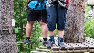Zwei Jungedliche klettern im Hochseilgarten: In Deutschland gelten rund zwei Millionen Kinder als übergewichtig, 800.000 von ihnen als adipös. Betroffene Kinder und Jugendliche kämpfen aber nicht nur mit ihren Pfunden, sondern auch mit Diskriminierung und Mobbing. Mehr Sportangebote, Ernährungskompetenz, Steuern auf Süßgetränke und Werbeverbote für ungesunde Kinderlebensmittel lauten einige Vorschläge zur Prävention.