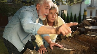 Ein älterer Mann und eine jüngere Frau stecken die Köpfe zusammen und er zeigt über einem Stapel Brennholz mit dem Finger auf etwas: Sprechen über andere hat ein schlechtes Image und wird schnell als Lästern kategorisiert. Zu Unrecht, finden manche Psychologen, denn das Reden über andere hat auch nützliche Funktionen in der Gesellschaft. Nur übertreiben darf man es nicht, sonst ist schnell auch der Tatbestand der Verleumdung erfüllt.