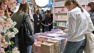 Mit Blumen geschmückter Messestand: Das neue Genre New Adult lockt auf der Frankfurter Buchmesse junge Leserinnen an. Booktoker und Bookfluencer besprechen auf Insta und Tiktok Bücher. Ihre Follower feiern sie dafür wie Popstars. Das bringt Verlagen neue Umsätze und mischt den Literaturbetrieb auf.