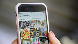 Zwei Hände mir rot lackierten Fingernägeln halten ein Smartphone, auf dem in einer Social-Media-App der Filter #booktook aktiviert ist: Booktoker und Bookfluencer besprechen auf Insta und Tiktok Bücher. Ihre Follower feiern sie dafür wie Popstars. Das bringt Verlagen neue Umsätze und mischt den Literaturbetrieb auf. 