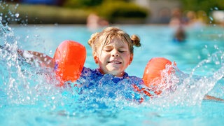 Kleines Mädchen mit Schwimmflügeln planscht im Pool: In den letzten Jahren steigt die Zahl der Grundschüler, die nicht schwimmen können. Die Gründe sind vielfältig: geschlossene Bäder während der Corona-Pandemie, zu wenige Schwimmlehrer und fehlende und marode Schwimmbäder. Warum ist guter Schwimmunterricht wichtig?