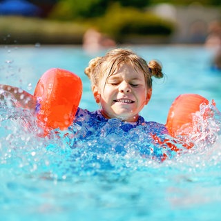 Kleines Mädchen mit Schwimmflügeln planscht im Pool: In den letzten Jahren steigt die Zahl der Grundschüler, die nicht schwimmen können. Die Gründe sind vielfältig: geschlossene Bäder während der Corona-Pandemie, zu wenige Schwimmlehrer und fehlende und marode Schwimmbäder. Warum ist guter Schwimmunterricht wichtig?