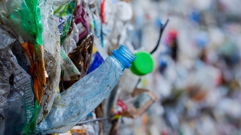 Gepresste und sortierte Verpackungsabfälle, eine Plastikflache ragt heraus: Die Erfindung von Plastik hat der Menschheit viele neue Möglichkeiten gegeben – doch heute bereitet es Probleme: Es verdreckt die Umwelt, ist schlecht fürs Klima und reichert sich als Mikroplastik in unseren Körpern an. Doch mit chemischen Tricks könnten Kunststoffe ökologisch viel besser werden.