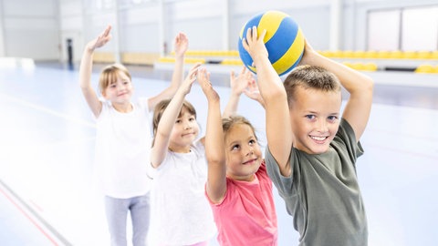 Kinder bei einem Ballspiel in einer Sporthalle: Zeitgemäßer Schulsport trainiert nicht nur verschiedene Sportarten, sondern auch soziale Fähigkeiten. Er motiviert Kinder und Jugendliche zur Bewegung.