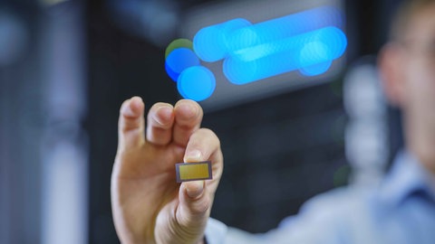 Eine Nahaufnahme einer Hand, die einen kleinen, goldenen photonischen Chip zwischen Daumen und Zeigefinger hält. Im Hintergrund sind unscharfe blaue Lichter zu sehen