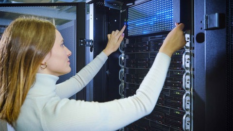 Eine Frau mit weißem Pullover interagiert mit einem photonischen Chip von Qant in einem Serverraum. Der Chip zeigt ein blaues Matrix-Display, das Licht aussendet und in einen Serverrack installiert ist. Die Photonen auf dem Chip ermöglichen energieeffizientes Computing ohne herkömmliche Kühlsysteme.