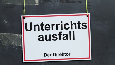 Ein weisses Schild mit rotem Rand und der Aufschrift "Unterrichtsausfall - Der Direktor" haengt schief an gelben Schnueren vor einer Tafel mit Resten von Klebeband.