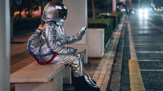 Eine als Astronaut verkleidete Person sitzt nachts an einer Bushaltestelle