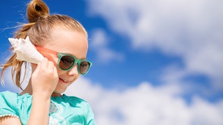 Ein Mädchen hält sich eine Meeresschnecke ans Ohr: Was wir in Muscheln oder Schnecken hören, ist nicht das Meeresrauschen. Es sind vielmehr die Geräusche aus der jeweils aktuellen Umgebung. Diese können sich in der Muschel verstärken.