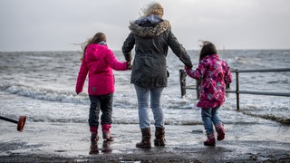 Eine Familie steht Hand in Hand bei Sturmflut an der Küste der Nordsee und schaut aufs Wasser: Bei Flut sind wir leichter und vom Erdmittelpunkt einen halben Meter weiter entfernt als bei Ebbe. Wie kann das sein?