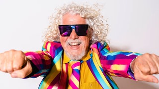 Älterer Mann mit grauem Haar und buntem Anzug lacht: Graue Haare sind eine optische Täuschung. Einzelne graue Haare gibt es gar nicht, denn sobald die Haarwurzel keine Pigmente mehr produziert, sind Haare farblos und erscheinen weiß. 