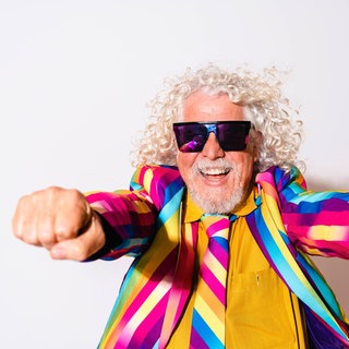 Älterer Mann mit grauem Haar und buntem Anzug lacht: Graue Haare sind eine optische Täuschung. Einzelne graue Haare gibt es gar nicht, denn sobald die Haarwurzel keine Pigmente mehr produziert, sind Haare farblos und erscheinen weiß. 