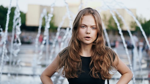 Eine junge Frau steht im schwarzen T-Shirt vor einem Springbrunnen: Ist ein Material dunkel, dann regen Lichtteilchen die Atome in diesem Material an bzw. bzw. versetzen sie in Schwingungen. Was bedeutet das?