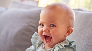 Säugling sitzt auf einer Couch und lacht:  Wenn ein Baby lacht, wenn man es kitzelt – und das tut es schon mit vier Monaten – ist das wirklich schon Humor? Oder nur ein Reflex oder einfach soziales Lachen? 