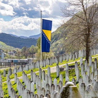 Eine bosnische Flagge weht auf Halbmast vor einer Reihe von Gräbern an der Gedenkstätte für den Völkermord von Srebrenica. Sie erinnert an die über 8.372 bosnischen Männer und Jungen, die von ihren Müttern, Schwestern und Töchtern getrennt und von bosnisch-serbischen Streitkräften Jugoslawiens getötet wurden.