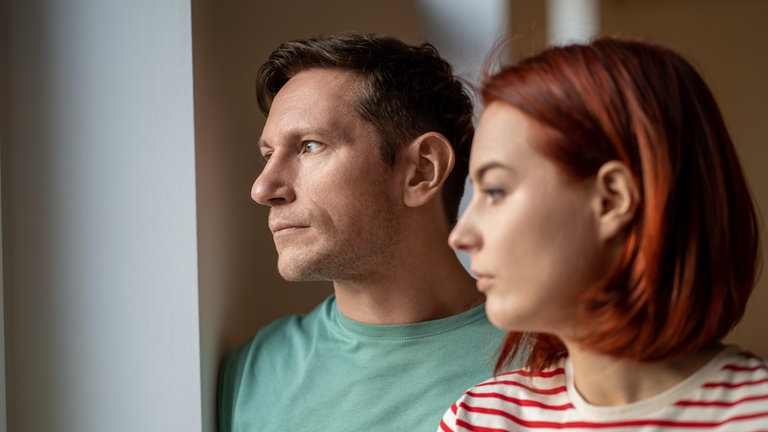 Ein Paar, Mann und Frau, stehen nebeneinander und schauen nachdenklich aus dem Fenster: Welcher Bindungstyp bin ich? Antworten liefert die Bindungstheorie, ein einflussreiches Konzept der Psychologie. Sie besagt, dass eine sichere Bindung zwischen Eltern und Kindern entscheidend dafür ist, ob wir als Erwachsene beziehungsfähig oder bindungsunfähig sind. 