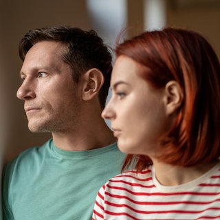 Ein Paar, Mann und Frau, stehen nebeneinander und schauen nachdenklich aus dem Fenster: Welcher Bindungstyp bin ich? Antworten liefert die Bindungstheorie, ein einflussreiches Konzept der Psychologie. Sie besagt, dass eine sichere Bindung zwischen Eltern und Kindern entscheidend dafür ist, ob wir als Erwachsene beziehungsfähig oder bindungsunfähig sind. 