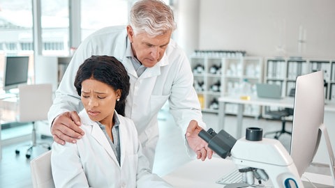 Grauhaariger Mann belästigt junge Frau. Sie arbeitet am Mikroskop, er legt ihr die Hand auf die Schulter: Es kann Doktoranden treffen und Spitzenforscherinnen. Ist die Wissenschaft besonders anfällig für Mobbing, Belästigung und Machtmissbrauch?