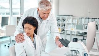Grauhaariger Mann belästigt junge Frau. Sie arbeitet am Mikroskop, er legt ihr die Hand auf die Schulter: Es kann Doktoranden treffen und Spitzenforscherinnen. Ist die Wissenschaft besonders anfällig für Mobbing, Belästigung und Machtmissbrauch?