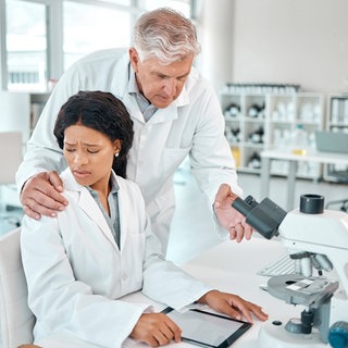 Grauhaariger Mann belästigt junge Frau. Sie arbeitet am Mikroskop, er legt ihr die Hand auf die Schulter: Es kann Doktoranden treffen und Spitzenforscherinnen. Ist die Wissenschaft besonders anfällig für Mobbing, Belästigung und Machtmissbrauch?