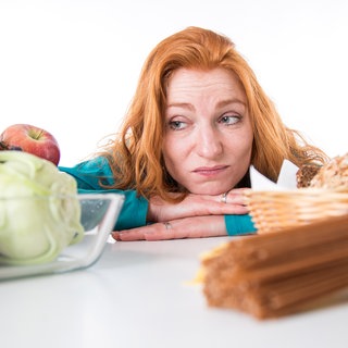 Eine Frau stützt Arme und Kopf auf einen Tisch und schaut missmutig auf eine Schale mit Ost und Gemüse: Wer raucht, sich nicht viel bewegt und schlecht ernährt, erhöht sein Risiko für viele schwere Erkrankungen. 