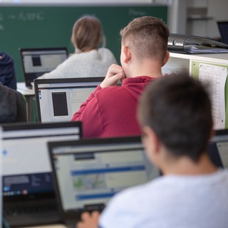 Schülerinnen und Schüler nehmen im Klassenzimmer einer 9. Klasse einer Gemeinschaftsschule mit Hilfe von Laptops und Tablets teil
