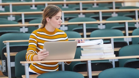Studentin mit Notebook und Büchern sitzt allein im leeren Hörsaal: Private Hochschulen boomen in Deutschland. Sie sind teuer, bieten aber denjenigen Chancen auf ein Studium, die an staatlichen Unis keine Möglichkeiten hätten.