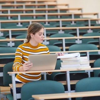Studentin mit Notebook und Büchern sitzt allein im leeren Hörsaal: Private Hochschulen boomen in Deutschland. Sie sind teuer, bieten aber denjenigen Chancen auf ein Studium, die an staatlichen Unis keine Möglichkeiten hätten.