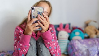 Symbolfoto zum Thema Handynutzung bei Kindern: Ein Mädchen sitzt in ihrem Bett und ist am Handy.
