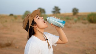 Junge Frau mit Hut trinkt Wasser aus einer Flasche in einer Wüstenregion: Unser Körper braucht nicht nur Wasser, sondern auch Salz. Und das Verhältnis zwischen beiden muss stimmen.