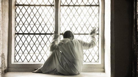 Frau in weißem Nachthemd am vergitterten Fenster einer Psychiatrie (Symbolbild): Bis in die 1970er-Jahre glichen Psychiatrien elenden Verwahranstalten: Patienten wurden ruhiggestellt, weggesperrt und unter menschenunwürdigen Bedingungen untergebracht.