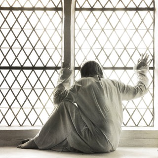 Frau in weißem Nachthemd am vergitterten Fenster einer Psychiatrie (Symbolbild): Bis in die 1970er-Jahre glichen Psychiatrien elenden Verwahranstalten: Patienten wurden ruhiggestellt, weggesperrt und unter menschenunwürdigen Bedingungen untergebracht.