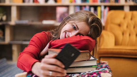 Studentin liegt mit dem Oberkörper auf einem Stapel Bücher und schaut froh in ihr Smartphone: Studierende scheitern oft an langen Texten. Klagen über die Leseschwäche an den Universitäten ziehen sich quer durch die Fächer. Ist das wirklich so schlimm? 