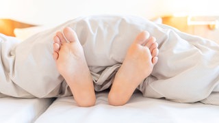 Zwei Füße schauen unter einer Bettdecke hervor: Kinder fallen im Schlaf gelegentlich aus dem Bett, weshalb man sie oft in Gitterbetten legt. Erwachsene dagegen brauchen das nicht. Warum ist das so? 