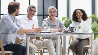 Ältere Kollegin inmitten ihres Teams: Lange galten die Wechseljahre als Privatsache. Doch es ändert sich etwas, auch im Job: Große Unternehmen wie Vodafone, Edeka und SAP ermöglichen flexiblere Arbeitszeiten und bieten Beratung. 