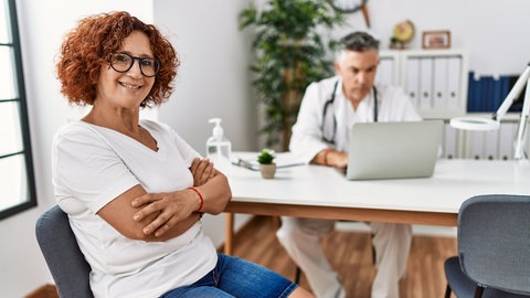 Frau sitzt zur Beratung in der Sprechstunde eines Arztes und lächelt: Über "die Wechseljahre" wissen Frauen oft wenig. Etwa dass das Östrogen schon ab Anfang 40 zu sinken beginnt. Und dass die Hormonersatztherapie gut wirkt und weitgehend sicher ist.