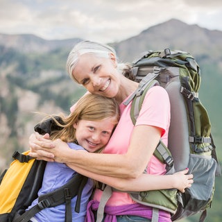 Großmutter und Enkelin mit Rucksäcken umarmen sich vor einem Bergpanorama: Wie wir schon als junge Menschen auf das Altwerden blicken, hat großen Einfluss darauf, wie wir selbst altern.