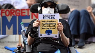 Demonstrantin und MECFS-Betroffene hält 2024 im Rollstuhl sitzend in Berlin ein Schild: "Wir fordern Aufklärung, Forschung, Versorgung". 1,5 Mio. Menschen in Deutschland haben Long Covid oder MECFS. Das erzeugt auch hohe Kosten für die Gesellschaft.