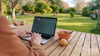 Digitaler Nomade arbeitet im Garten am Laptop: Manche Kommunen reagieren mit kreativen Strategien auf den Leerstand – mit Probewohnen, Zuschüssen für Baugrundstücke, gezieltem Standortmarketing oder der Errichtung digitaler Nomadendörfer.